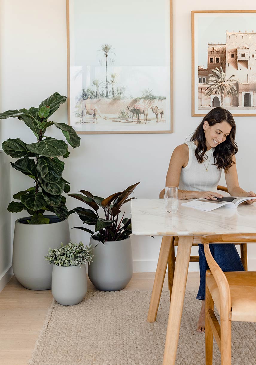 Three grey pots with indoor plants are styled behind a smiling woman sitting at a dining table browsing a design magazine..