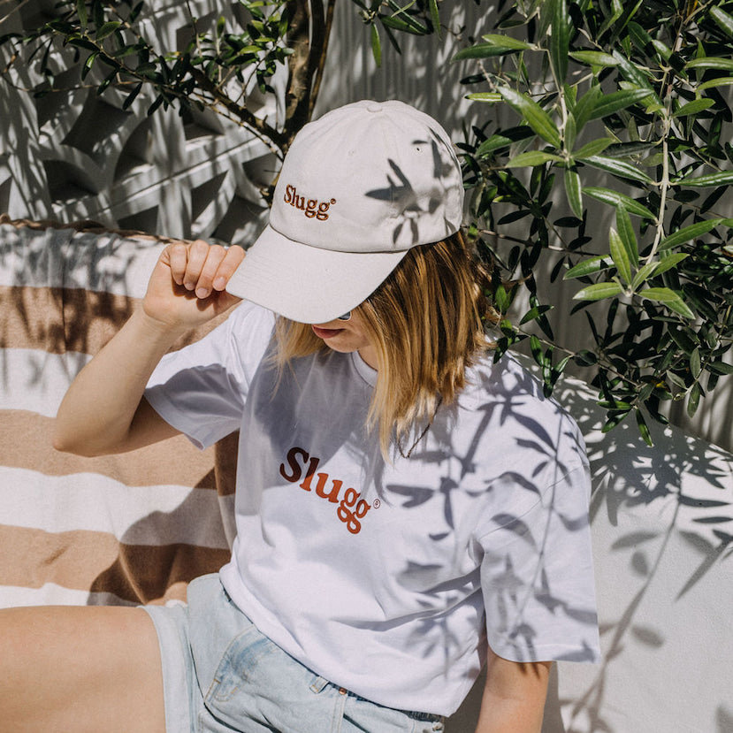A plant enthusiast sits in the shade of an Olive Tree wearing a Slugg branded  taupe cap and white coloured t-shirt. 